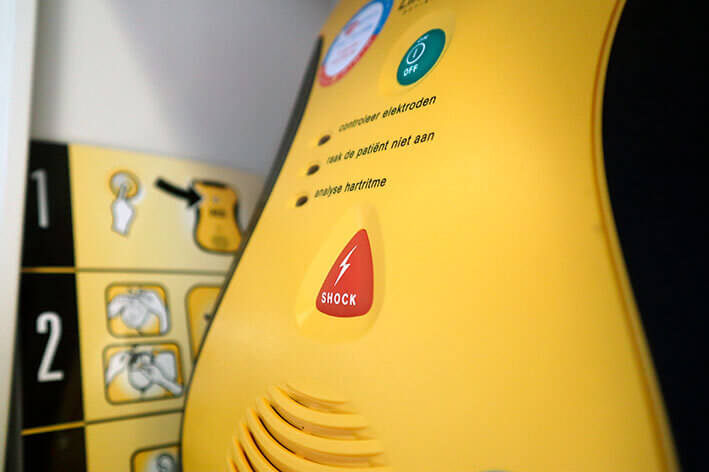 Close-up van een gele automatische externe defibrillator (AED) met een rode Schok-knop en gedeeltelijk zichtbare instructies op de achtergrond. Wat als je geen AED in je bedrijfspand hebt hangen? Zorg voor veiligheid door er een toegankelijk te hebben.