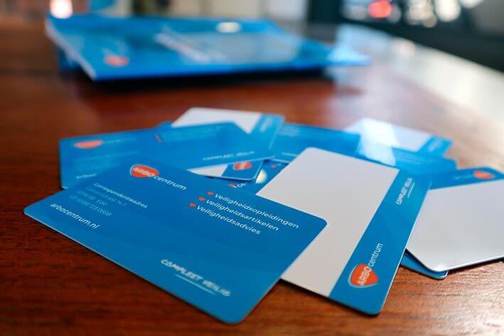 Blauwe en witte kaarten verspreid over een houten tafel, geaccentueerd met oranje en witte tekst, sommige onthullen gedeeltelijke ontwerpen of lege ruimtes. De wazige achtergrond hint naar vijf tips om je voor te bereiden op je VCA-examen, omringd door extra blauwe objecten in het zicht.