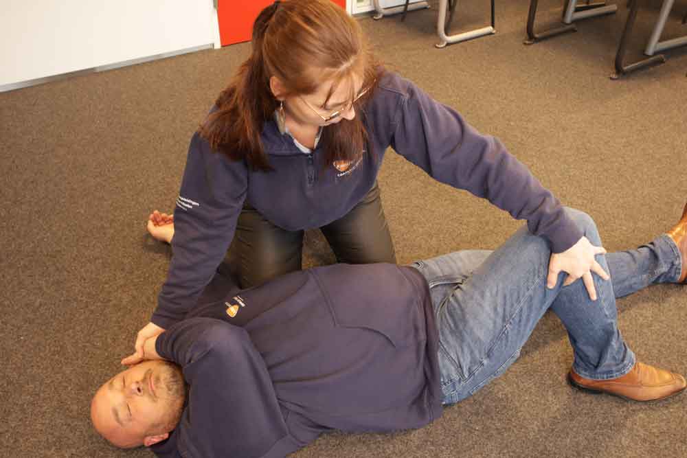 Een persoon in een donkerblauw sweatshirt wordt in de stabiele zijligging op de grond geplaatst door een ander persoon die een bril en een soortgelijk sweatshirt draagt. De assisterende persoon corrigeert de armpositie van de persoon op de grond—zo herken je een beroerte.