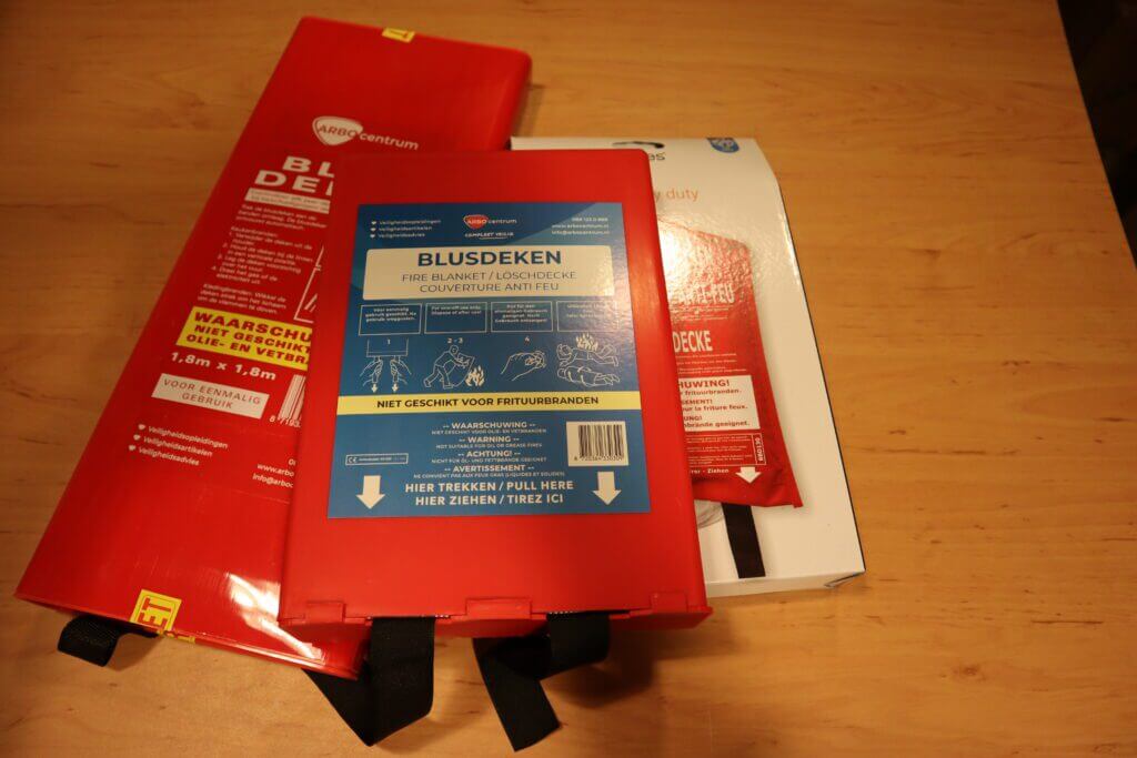 Een rode branddekenverpakking met labels in verschillende talen rust op een houten oppervlak. Met veiligheidspictogrammen en beknopte instructies, waaronder 5 stappen om een blusdeken te gebruiken bij mensen, heeft het twee zwarte treklipjes aan de onderkant voor snelle toegang.