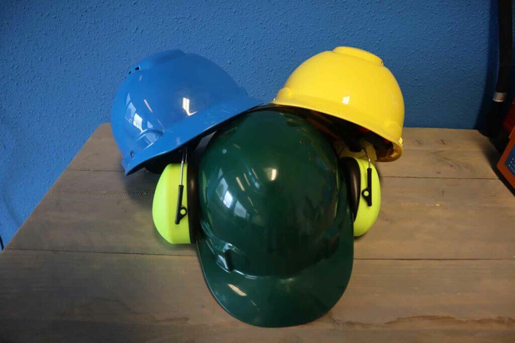 Drie bouwhelmen in blauw, geel en groen zijn gestapeld op een houten oppervlak. De groene helm is voorzien van gehoorbescherming. Tegen de contrasterende blauwe muur stellen ze de vraag: ‘Moet ik mijn VCA basis behalen voordat ik mijn VCA VOL diploma wil halen?.