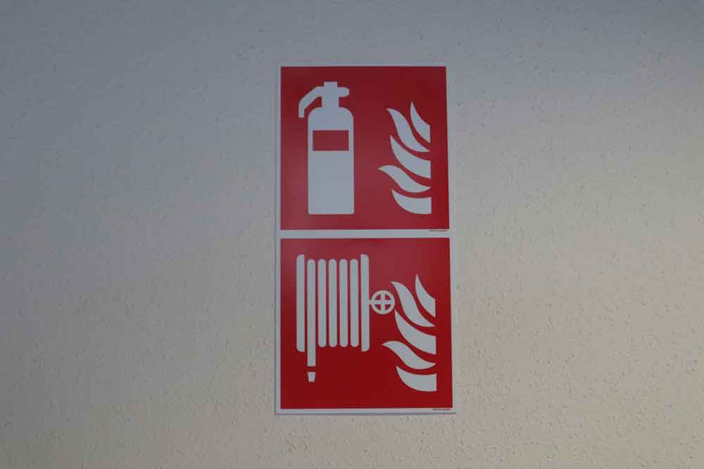Twee rood-witte brandveiligheidsborden sieren de muur. Het bovenste bord toont een brandblusser naast vlammen, met de stappen die u moet nemen in geval van brand. Daaronder toont een ander bord een brandslang naast vlammen als onderdeel van de richtlijnen deze stappen volg je indien er een brand uitbreekt.