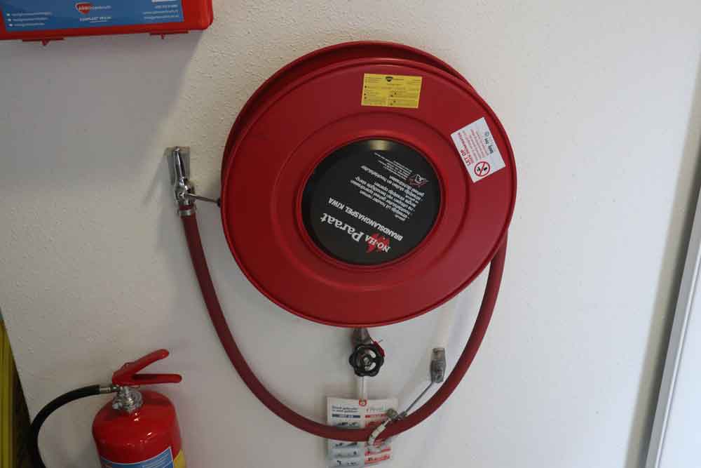Een rode brandslanghaspel, compleet met een opgerolde slang en spuitmond, is gemonteerd op een ongerepte witte muur. Daaronder staat een rode brandblusser op de vloer. Labels en instructies zijn duidelijk zichtbaar op zowel de haspel als de blusser—een brandslanghaspel of een brandblusser?.