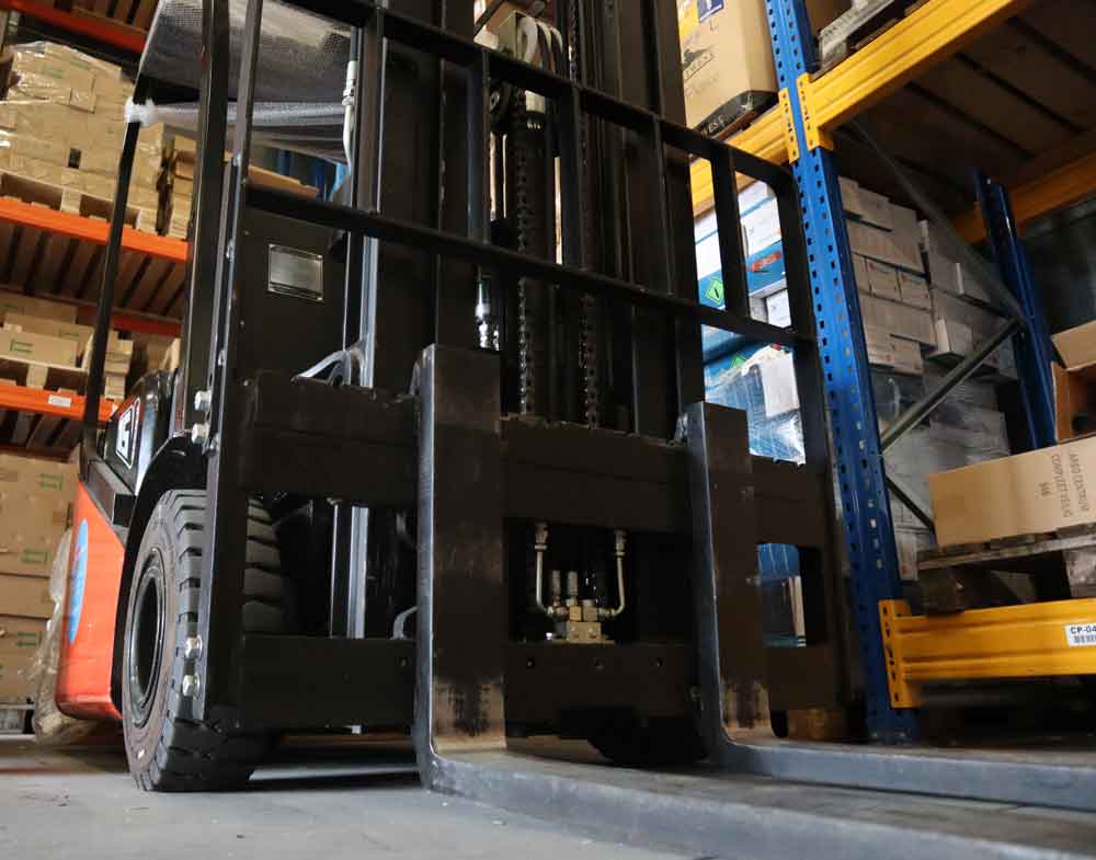 Close-up van een vorkheftruck in een magazijn, gepositioneerd bij planken vol met kartonnen dozen. De vorken van de vorkheftruck zijn verlaagd en de wielen zijn zichtbaar - een herinnering aan de 3 redenen om je heftruckcertificaat te beperkingen. De omringende metalen planken hebben oranje en blauwe componenten.