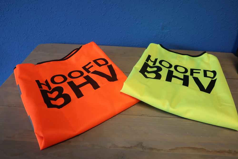 Twee opgevouwen vesten liggen op een houten tafel tegen een blauwe muur. Het ene vest is feloranje, het andere neongeel, beide met HOOFD BHV in grote zwarte letters op de voorkant. Om deze redenen heb je een hoofd BHV: veiligheid en zichtbaarheid staan voorop.