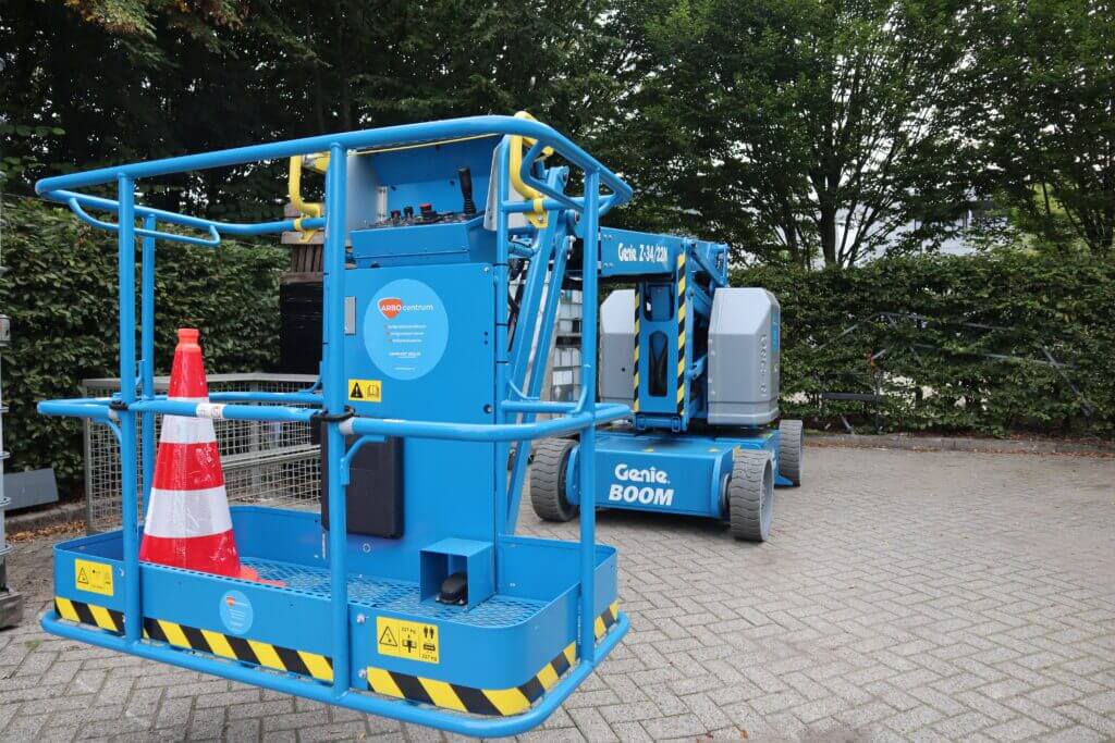 Een blauwe Genie hoogwerker rust op een geplaveid terrein omringd door bomen en heggen, met een oranje en witte verkeerskegel op het platform. De machine, versierd met gele waarschuwingslabels, wacht terwijl hierom laat je jouw medewerkers een IPAF cursus volgen om de veiligheid te waarborgen.