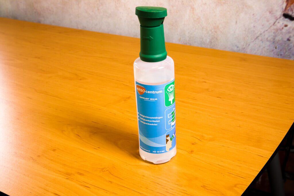 Een fles met een groene dop en een blauw etiket, dat slim suggereert Dit kun je met een oogdouche voorkomen, staat op een houten tafel. Het etiket bevat wat tekst en afbeeldingen, tegen de achtergrond van een getextureerde muur.