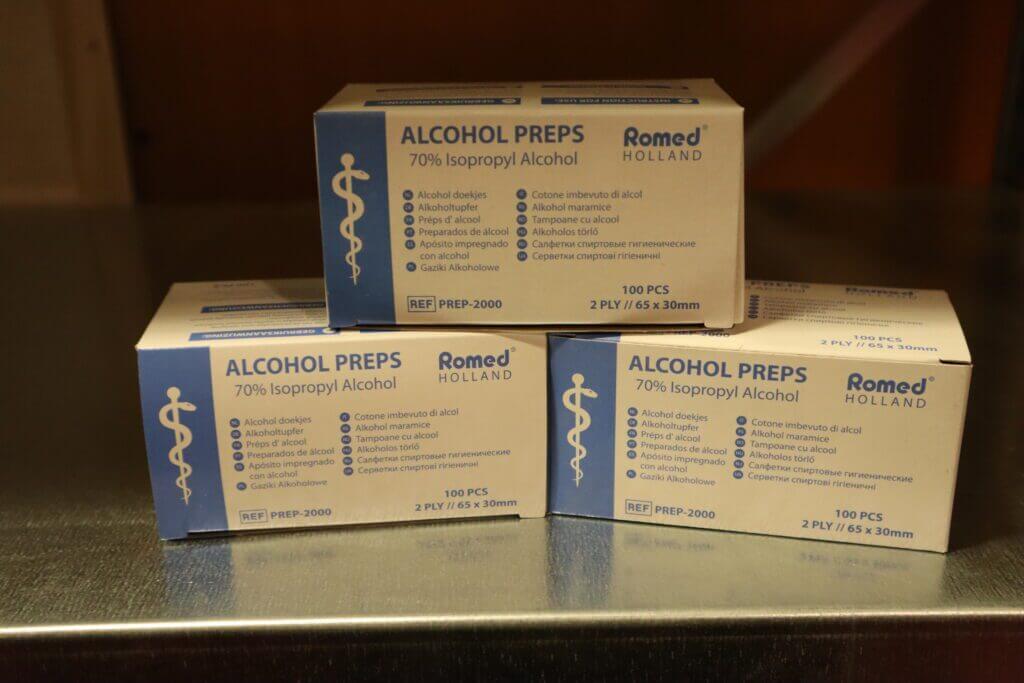 Drie dozen Romed Holland alcohol prep pads zijn netjes gestapeld op een metalen oppervlak. Elke doos, ideaal om de voordelen van alcoholdoekjes op het kantoor te benadrukken, bevat 100 stuks 2-laags 65 x 30 mm pads met 70% isopropylalcohol. De verpakking is blauw en wit, met medische symbolen.