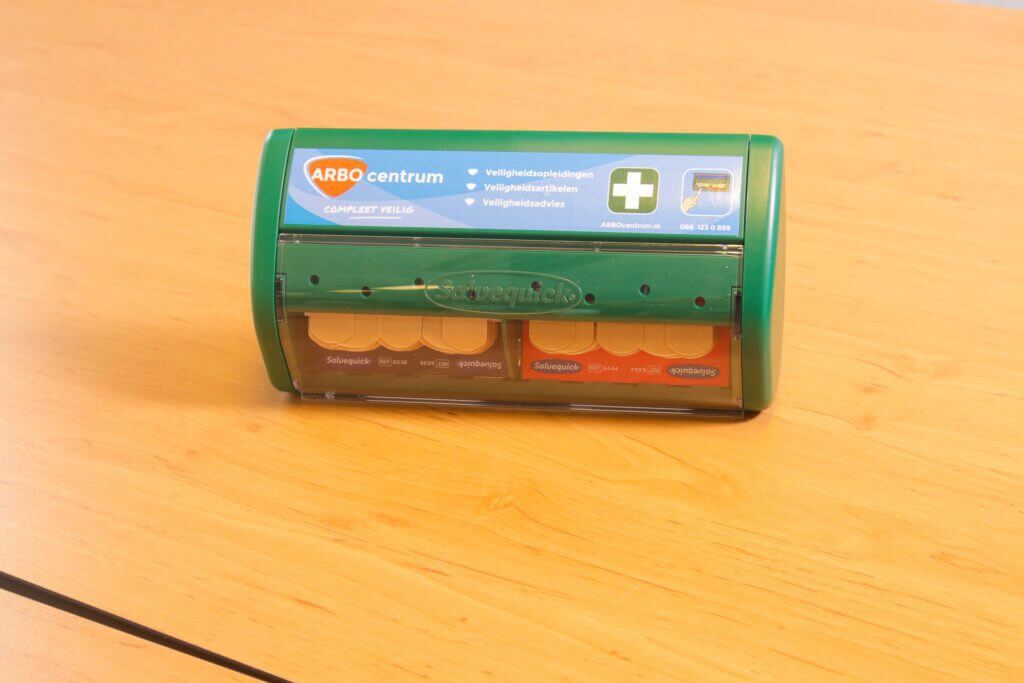Een groene en transparante EHBO-dispenser met het label ARBO centrum staat op een houten oppervlak. Ideaal voor horecagebruik, het bevat verschillende soorten pleisters netjes georganiseerd in compartimenten.