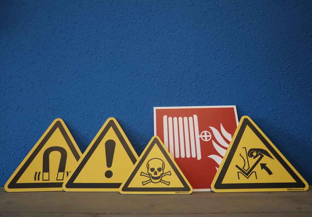 Deze 5 veiligheidspictogrammen zijn aan te raden in een bedrijfspand: vier gele, driehoekige waarschuwingsborden met symbolen zoals een magneet, uitroepteken, doodshoofd en hefpersoon. Achter hen staat een rood bord met radiator en vlam, allemaal tegen een blauwe muur geplaatst.