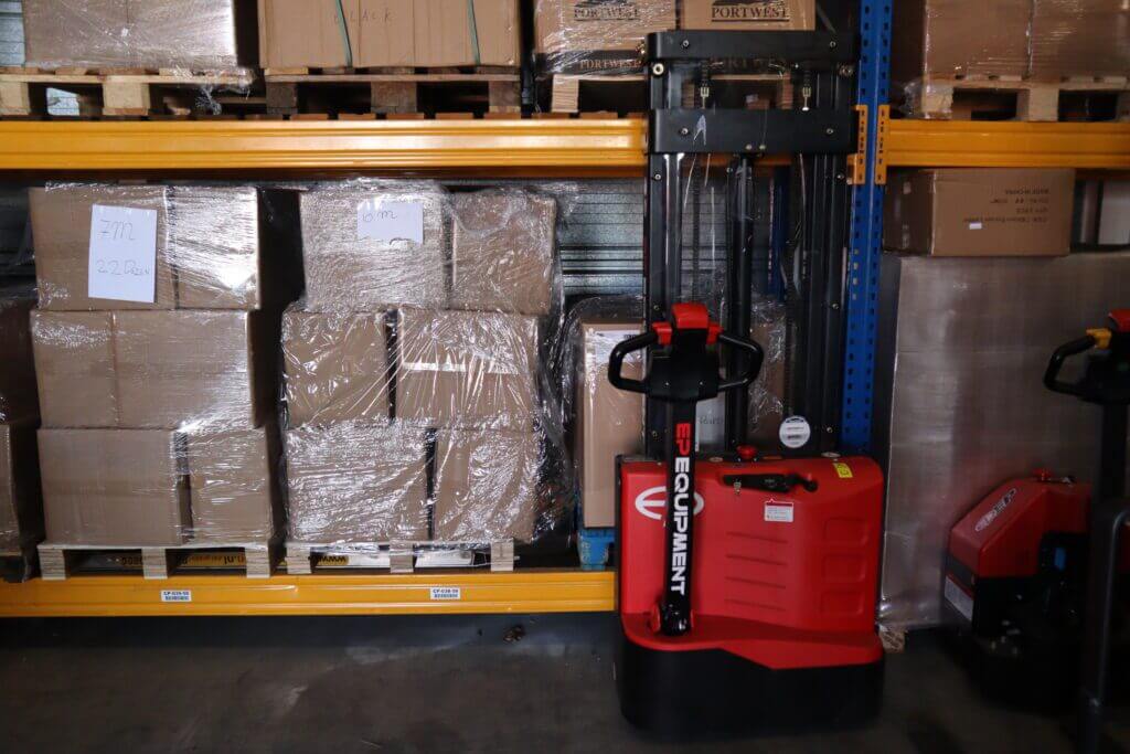 Een rode palletwagen staat geparkeerd in een magazijn naast planken met gestapelde kartonnen dozen. Sommige dozen zijn in plastic verpakt en op een paar zijn handgeschreven notities zichtbaar. De gele en blauwe planken markeren het gebied en illustreren verschillen die vergelijkbaar zijn met die tussen een EPT en een stapelmachine.