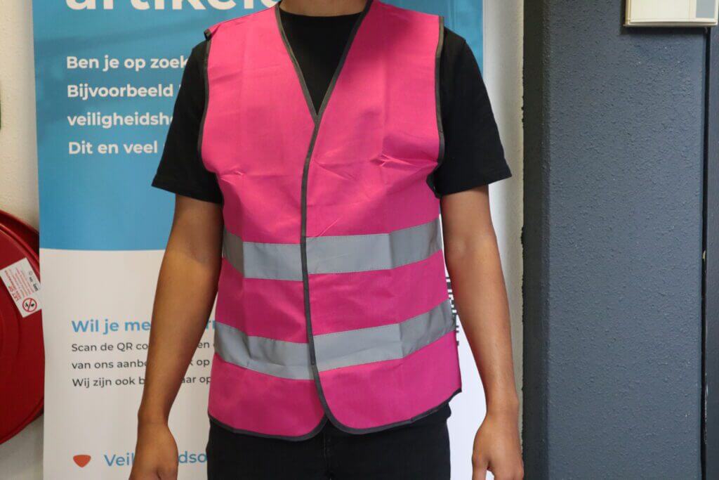 Een persoon draagt een roze veiligheidsvest met reflecterende zilveren strepen en staat voor een bord met daarop één van de 5 situaties waarbij je veiligheidshesjes nodig hebt. De persoon is zichtbaar van schouders tot middel en op de achtergrond is tekst en een rood brandalarm op de muur te zien.