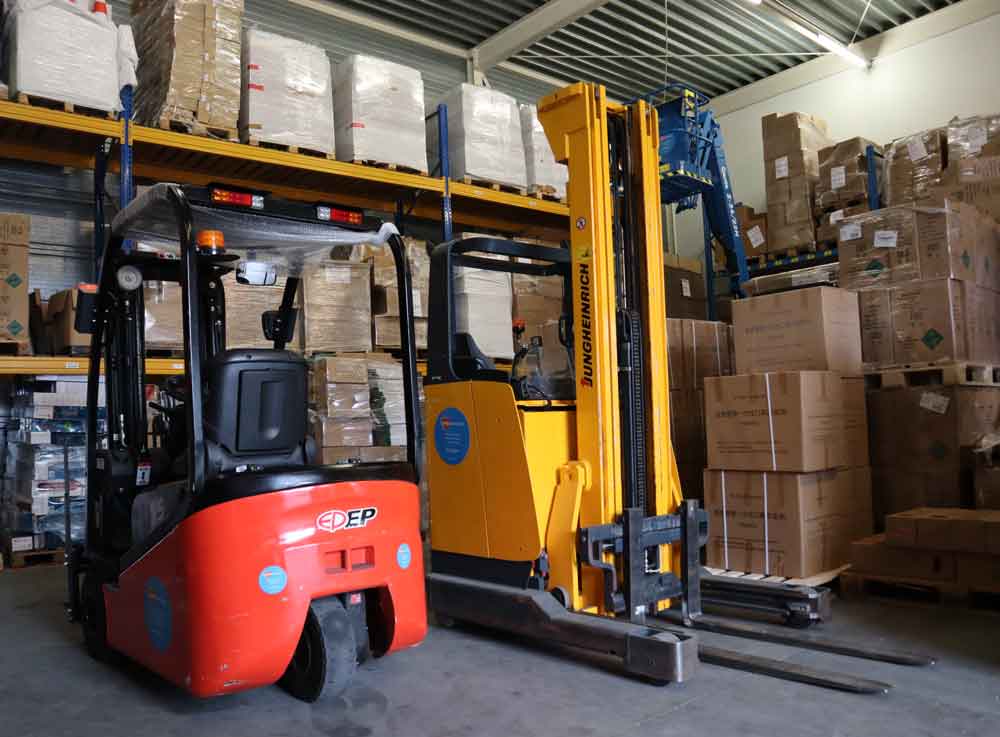 Twee vorkheftrucks, een rode en een gele, staan geparkeerd in een magazijn vol met stapels kartonnen dozen en pallets. Planken staan vol met grote ingepakte pakketten onder metalen balken en lampen, klaar voor de De heftruck en reachtruck cursus op 1 dag om de operationele efficiƫntie te maximaliseren.
