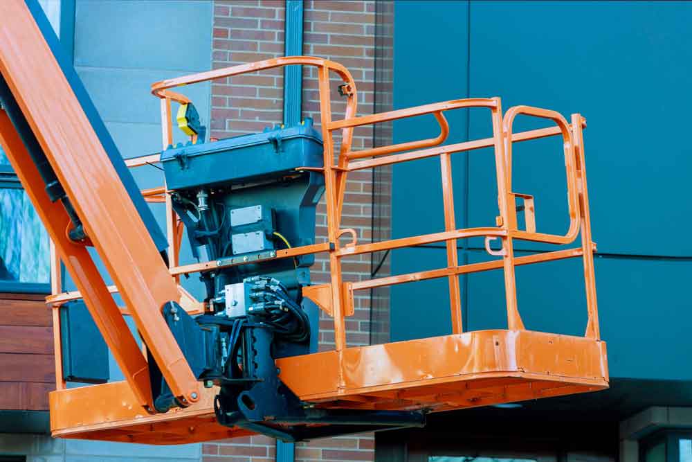 Een oranje bouwliftplatform is gepositioneerd bij een bakstenen en blauwgroene gevel van een gebouw. De lift is leeg, met mechanische componenten zichtbaar onder de reling, wat situaties benadrukt waarin je een hoogwerker gebruikt voor bouw- of onderhoudswerkzaamheden aan de buitenkant van een gebouw.