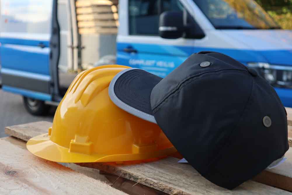 Een gele helm en een zwarte baseballpet, die het verschil tussen een stootpet en een veiligheidshelm illustreren, rusten op een houten oppervlak. Op de achtergrond is een blauw-witte bestelwagen met een open deur zichtbaar.