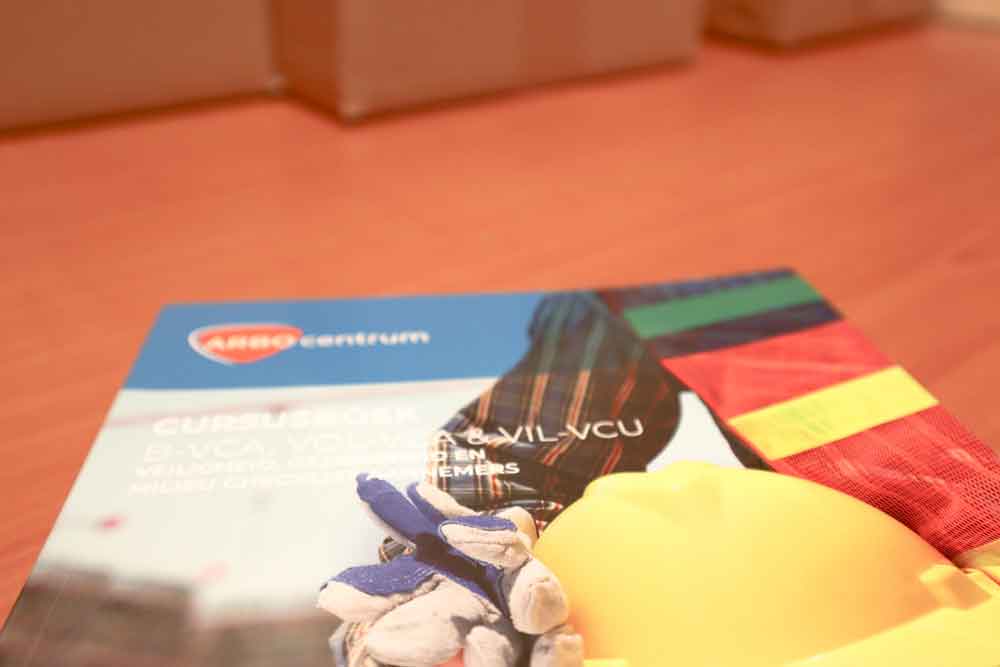 Een close-up van een brochure op een houten tafel toont een gehandschoende hand, een gele helm en felle strepen. Het merklogo centrum staat in de linkerbovenhoek. In deze talen is het VCA examen beschikbaar subtiel opgemerkt terwijl kartonnen dozen vervagen op de achtergrond.