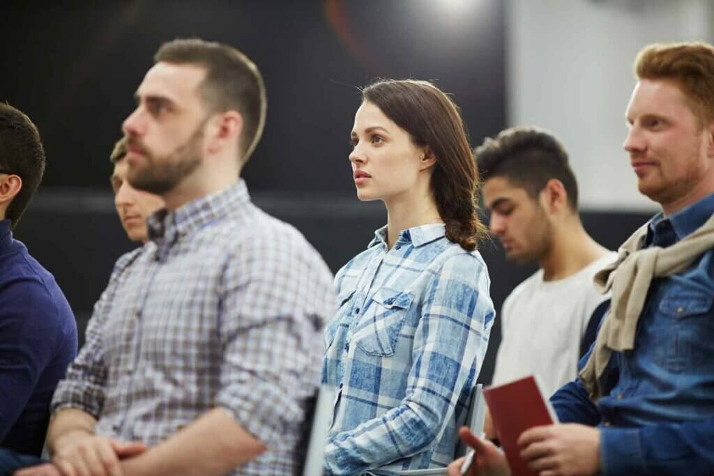 Een groep mensen zit aandachtig in een lezing en laat de voordelen van groepstraining zien. Gekleed in casual kleding, valt er eentje op in een blauw geruit overhemd. Notities en notitieboekjes zijn zichtbaar, wat de actieve betrokkenheid benadrukt, terwijl de achtergrond wazig blijft.
