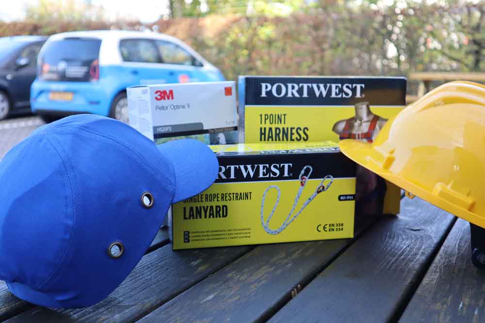 Veiligheidsmateriaal dat op een tafel wordt tentoongesteld, omvat een blauwe pet, een gele helm en een stofmaskerdoos. Twee zwarte en gele Portwest-dozen bevatten een 1-puntsgordel en een lanyard, essentieel voor veilig werken langs de weg. Een kleine blauwe auto staat op de achtergrond geparkeerd.