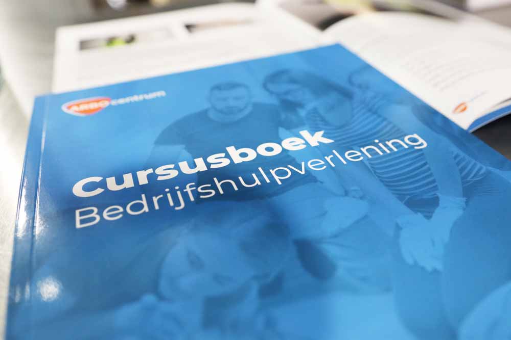 Een close-up van een blauw boek met de titel Cursusboek Bedrijfshulpverlening ligt op tafel en trekt de aandacht. Op de achtergrond biedt een open boek een inkijkje in 4 redenen om je BHV diploma te behalen, wat de nieuwsgierigheid prikkelt en het belang van EHBO-training onderstreept.
