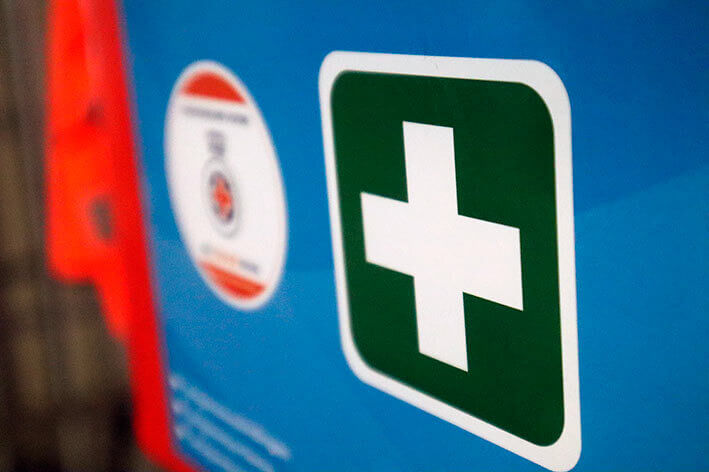 Een close-up van een bord over hoe je het nieuwe jaar veilig kunt omarmen, met een groen vierkant en wit kruis op een blauwe achtergrond, wat duidt op eerste hulp. Aan de linkerkant is een cirkelvormig logo met tekst en symbool gedeeltelijk zichtbaar.