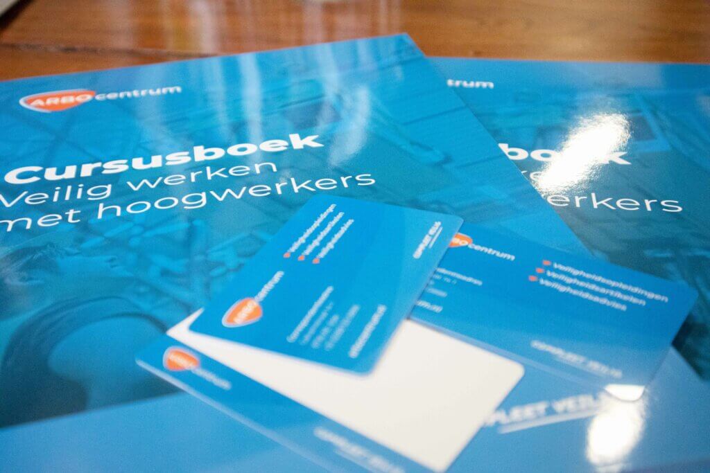 Close-up van een Nederlands cursusboek met de titel Cursusboek Veilig werken met hoogwerkers met verschillende blauwe kaarten bovenaan, met 3 tips om snel je hoogwerker certificaat te behalen. Op de achtergrond staat een ander soortgelijk boek.