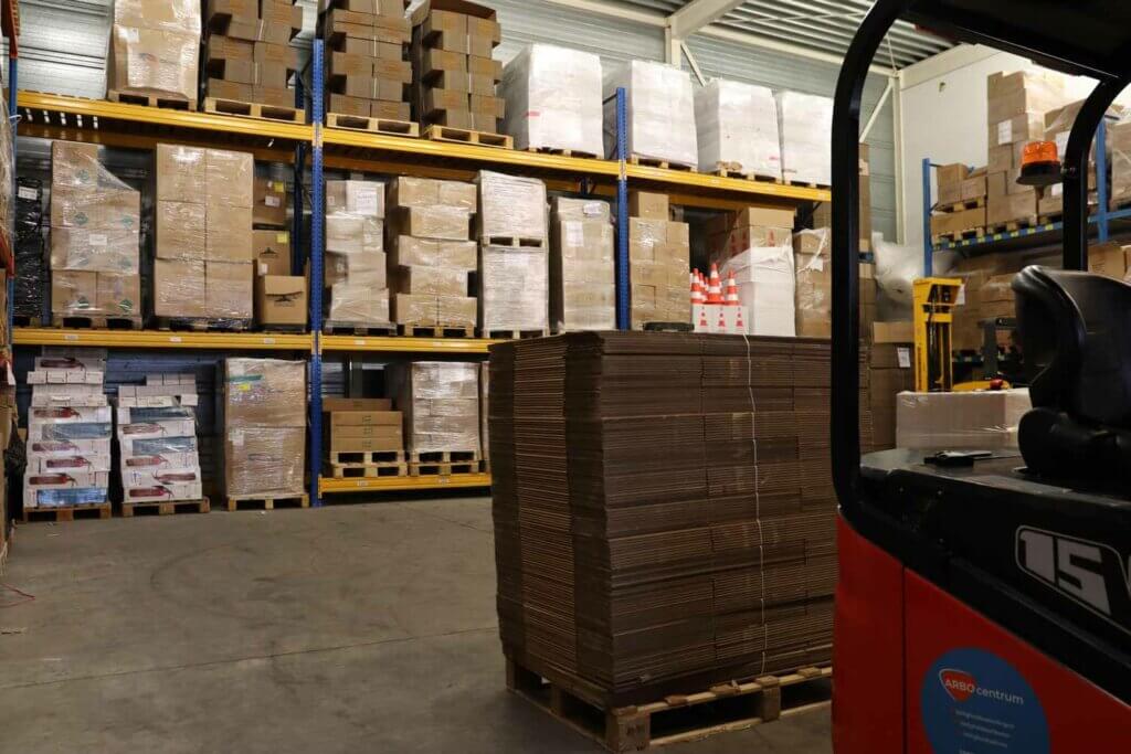 Het magazijninterieur gonst van activiteit, rijen schappen vol met dozen en pallets. Op de voorgrond is gedeeltelijk een vorkheftruck zichtbaar, een knipoog naar de 5 tips om veilig in een magazijn te werken. Industriële verlichting accentueert de minutieus georganiseerde inventaris.