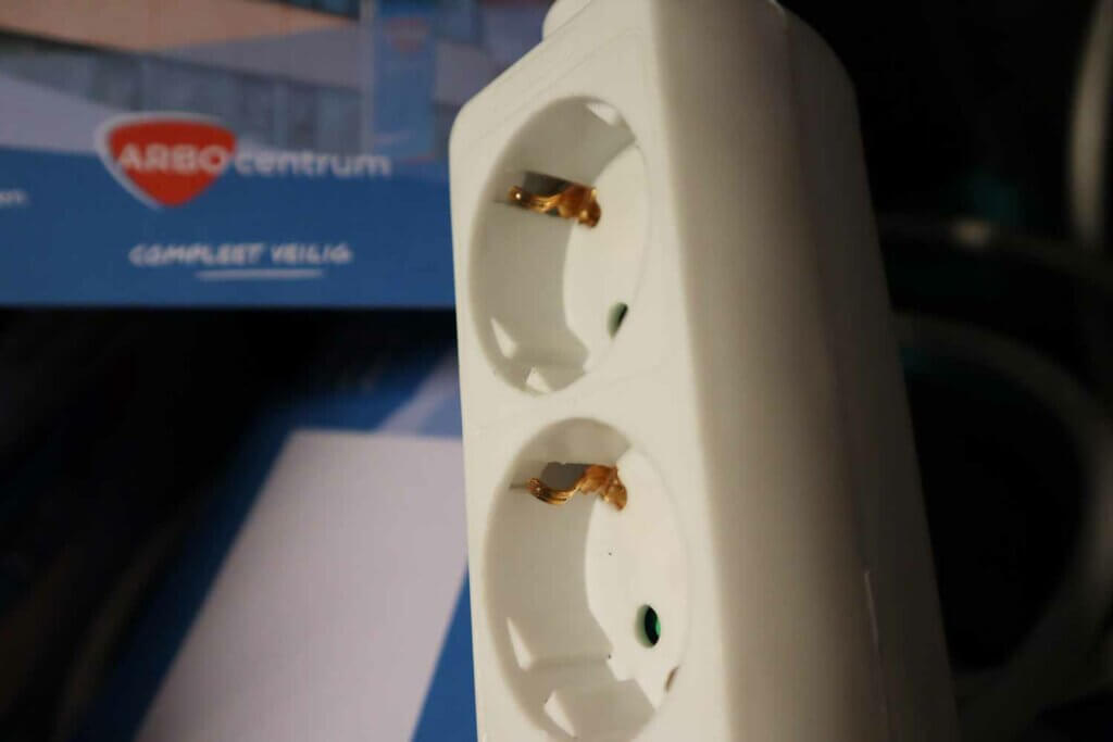 Close-up van een witte stekkerdoos met twee stopcontacten, die veiligheid in lijn met de voordelen van NEN 3140 VP laat zien. De achtergrond toont een wazig blauw-wit document met ARBOcentrum en een gedeeltelijk zichtbaar rood logo.