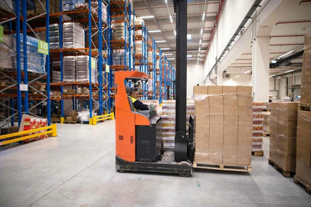 Een werknemer bedient een oranje vorkheftruck en manoeuvreert vakkundig een pallet met dozen door een groot magazijn met hoge blauwe en oranje planken vol goederen. De ruimte is goed verlicht met een hoog plafond, wat het belang van het behalen van uw reachtruckcertificaat benadrukt.