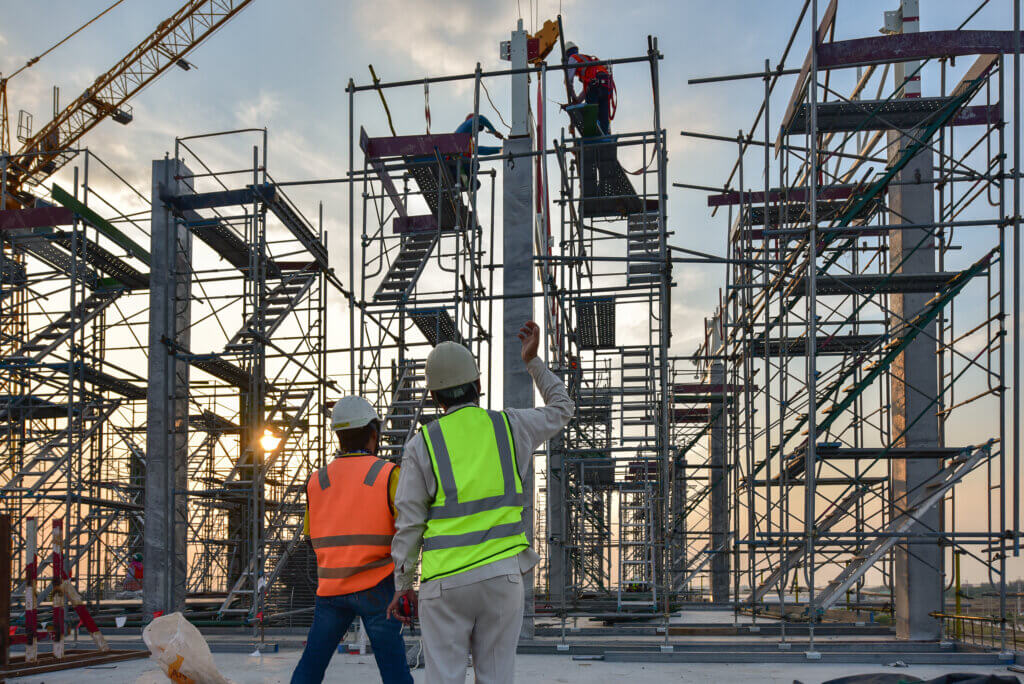Op een drukke bouwplaats observeren twee arbeiders in veiligheidsvesten en helmen de steiger. De een wijst omhoog terwijl de ander de metalen constructie beklimt, wat een van de 3 redenen om een rolsteiger certificaat te halen illustreert. Op de achtergrond doemt een hijskraan op tegen een bewolkte lucht.
