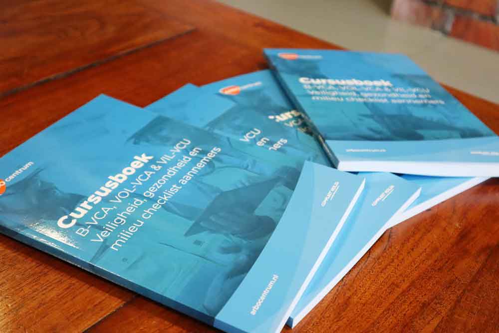 Verschillende blauw-blauwe Cursusboek trainingshandleidingen liggen op een houten tafel. De Nederlandse tekst behandelt onderwerpen over veiligheid, gezondheid en milieu. Een website-URL onderaan begeleidt lezers bij het voorbereiden op een VCA-examen.