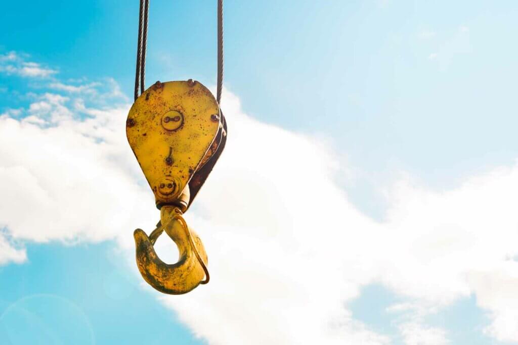 Close-up van een gele kraanhaak die tegen een helderblauwe lucht met verspreide wolken hangt. Het verweerde metaal suggereert veelvuldig gebruik, wat het belang van het nut van een diploma veilig hijsen en aanslaan van lasten onderstreept. Zonlicht benadrukt de haak en werpt een schaduw op één kant.
