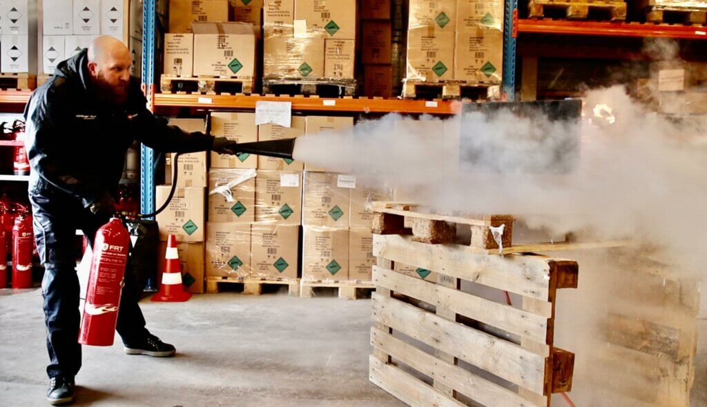 Tussen stapels kartonnen dozen en veiligheidskegels demonstreert een persoon één van de 3 voordelen van de BHV herhalingscursus door vakkundig een brandblusser te gebruiken om een brand op een houten pallet in het magazijn te blussen.