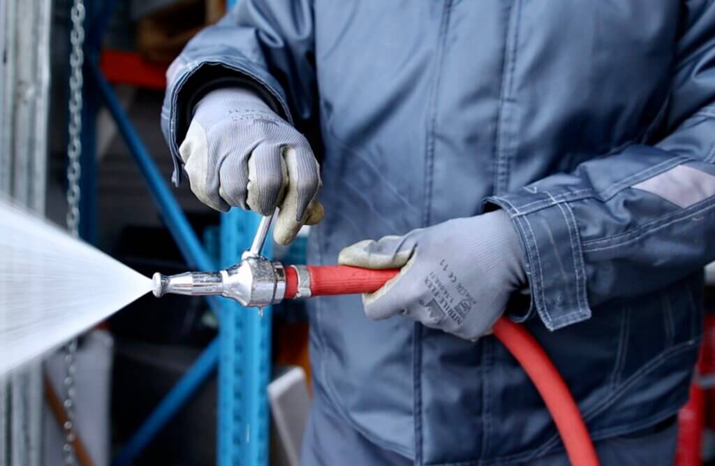 Close-up van een persoon in een blauw beschermend pak en handschoenen die een hogedrukwaterslang bedient, wat de vaardigheden laat zien die je kunt verwerven als je een BHV-cursus volgt. Het water spuit krachtig tegen een industriële achtergrond van metalen constructies.