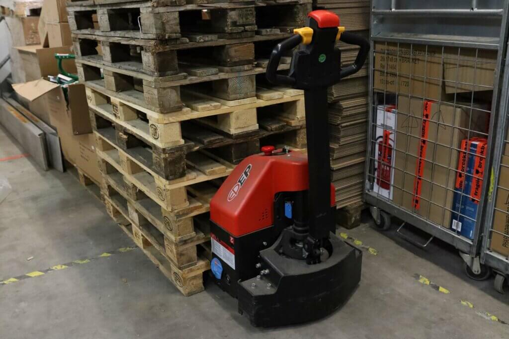 Een rode elektrische palletwagen, die de voordelen van een EPT laat zien, staat geparkeerd naast een stapel houten pallets in een magazijn. In de buurt zijn kartonnen dozen en een metalen kar vol spullen zichtbaar. De vloer is gemarkeerd met gele tape.