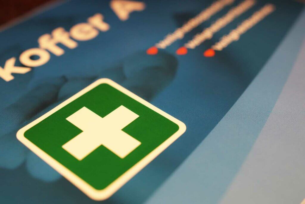 Close-up van een groen vierkant embleem met een wit kruis, dat medische hulp oproept, op een blauwe achtergrond. Gedeeltelijke tekst Koffer A is zichtbaar te midden van de wazige achtergrond en drie rode bullet points, wat duidt op 3 situaties waarin je eerste hulp moet worden genoemd.