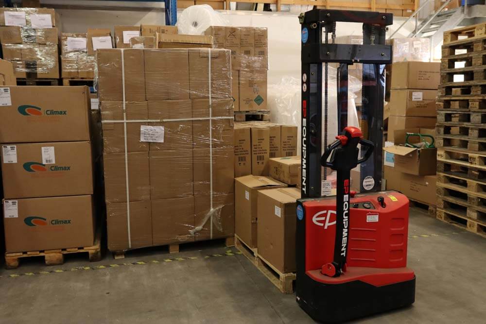 Een rode elektrische palletstapelaar staat geparkeerd in een gangpad in een magazijn en toont de voordelen van een stapelaar tussen rijen gestapelde kartonnen dozen en houten pallets. Planken, vol met nog meer dozen, strekken zich uit naar de achtergrond over de gladde betonnen vloer.