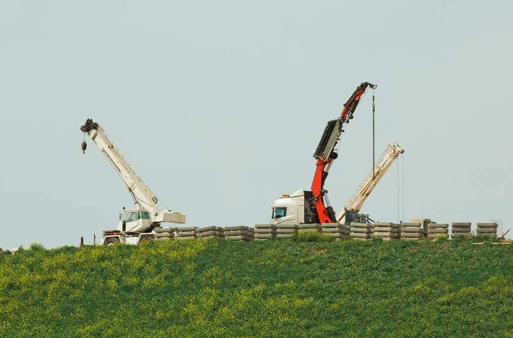 Twee grote kranen staan op een met gras begroeide heuvel, omringd door stapels banden. Met hun armen uitgestrekt tegen een heldere hemel benadrukken ze het belang van vakkundige bediening, slechts een van de 3 redenen om het certificaat chauffeur autolaadkraan te benutten in dergelijke bouw- of industriële omgevingen.