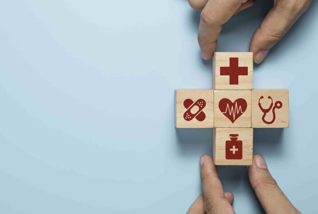 Handen rangschikken houten blokken met medische symbolen: een kruis, pil, hart met hartslag, stethoscoop en een medicijnfles. Deze vertegenwoordigen de fundamenten van gezondheidszorg en veiligheid, net als de 4 redenen om een BHV plan te maken voor elke organisatie. De blokken vormen een plusteken op een lichtblauwe achtergrond.