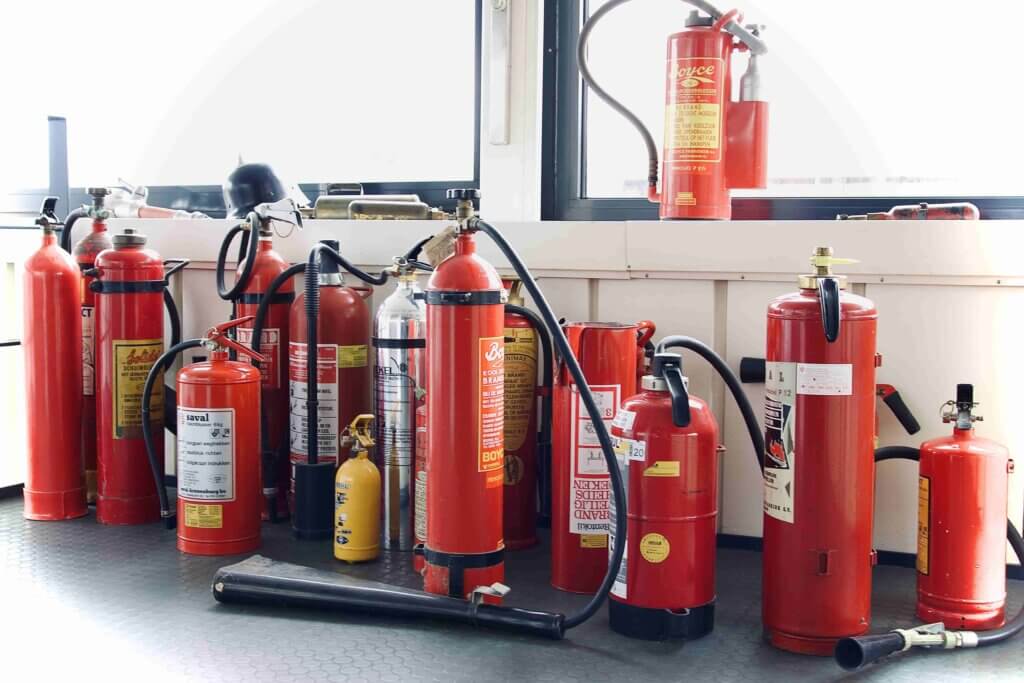 Een verzameling van verschillende rode brandblussers is netjes gerangschikt op de vloer bij een raam. Sommige met slangen, ze variëren in grootte van klein tot groot, en vormen een essentiële veiligheidsuitrusting display of opslagruimte. Dit zijn de nadelen van een vlot BHV-diploma: cruciale paraatheid kan in gevaar komen.