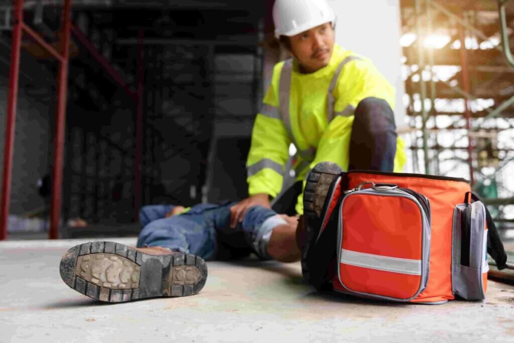 Een bouwvakker in een fluorescerend hesje en een helm verzorgt een gewonde collega die op de grond ligt. De arbeider houdt het been van de gewonde vast. Een feloranje EHBO-doos staat op de voorgrond.