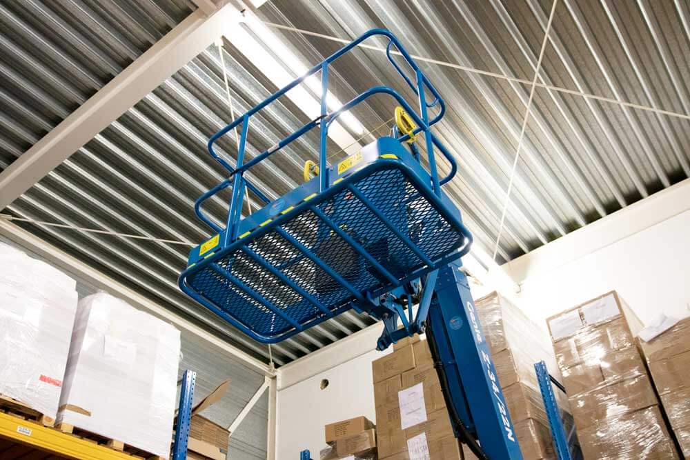 Een blauw schaarliftplatform staat opgetild in een magazijn met een gegolfd metalen plafond, omringd door gestapelde kartonnen dozen en pallets. De lift, essentieel voor het volgen van 4 tips voor veilig werken op hoogte, is gepositioneerd tussen de opslagplanken onder felle plafondlampen.