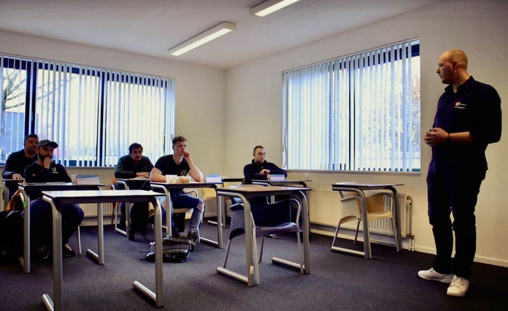 Een man staat vooraan in een klaslokaal en bespreekt de voordelen van een VCA-groepscursus met zes zittende studenten. Bureaus en stoelen zijn netjes opgesteld en ramen met gedeeltelijk open jaloezieën laten zachte stromen natuurlijk licht de kamer vullen.