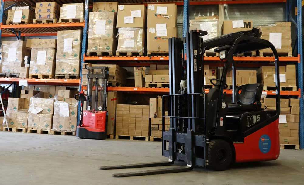 Een magazijninterieur toont gestapelde kartonnen dozen op metalen planken. Er zijn twee vorkheftrucks aanwezig, die klassieke scenarios illustreren waarin je ze nodig hebt: een rood-zwarte op de voorgrond en een kleinere rode op de achtergrond, geparkeerd bij de planken.