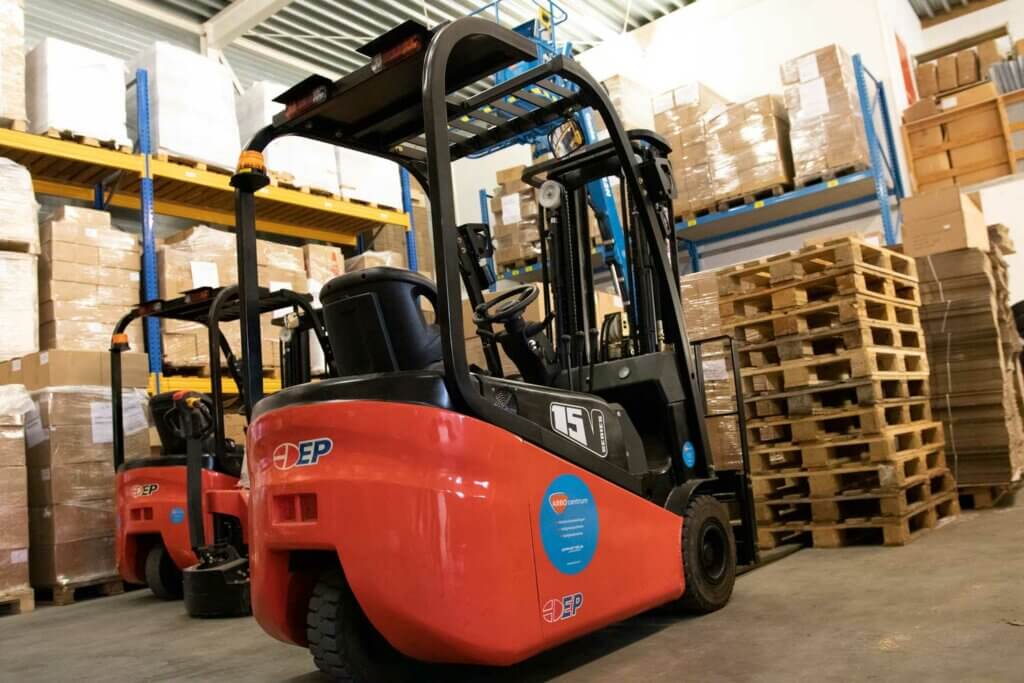 Een rode elektrische vorkheftruck staat geparkeerd in een loods, omringd door stapels dozen en houten pallets. Een andere soortgelijke vorkheftruck is zichtbaar op de achtergrond, wat het belang van een goede bediening benadrukt, want de nadelen van het rijden zonder je heftruck certificaat kunnen niet worden genegeerd. De ruimte is voorzien van metalen schappen en een dak van gegolfd metaal.