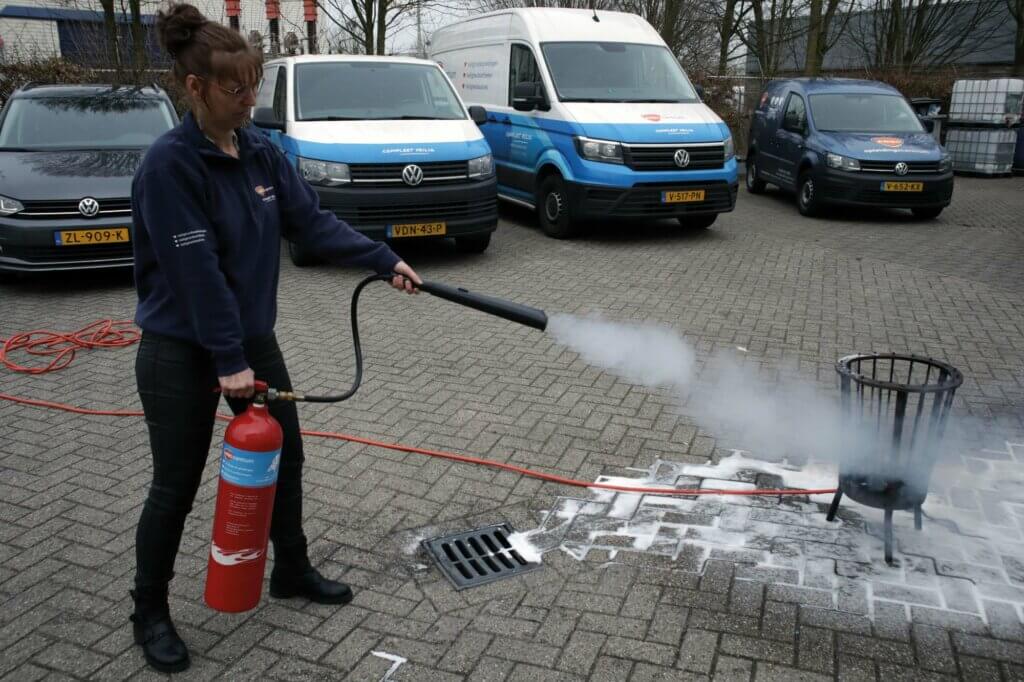 Een persoon in een blauw shirt laat zien hoe nuttig een BHV-opfriscursus is. Hij gebruikt hiervoor een rode brandblusser om een kleine brand in een metalen bak buiten te blussen. Achter hem staan geparkeerde busjes en autos op het verharde oppervlak.