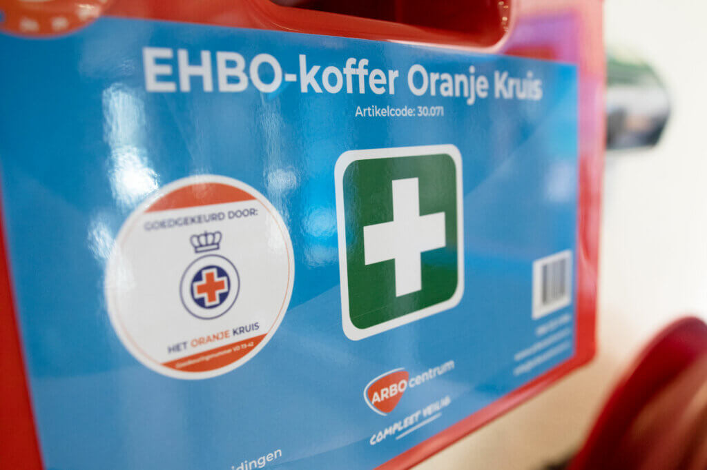 Close-up van een rode EHBO-doos met het opschrift EHBO-koffer Oranje Kruis met een wit kruis op een groene achtergrond. Het benadrukt certificeringsinformatie, logos en benadrukt de nadelen van het niet laten keuren van je EHBO-koffers voor veiligheidsborging.