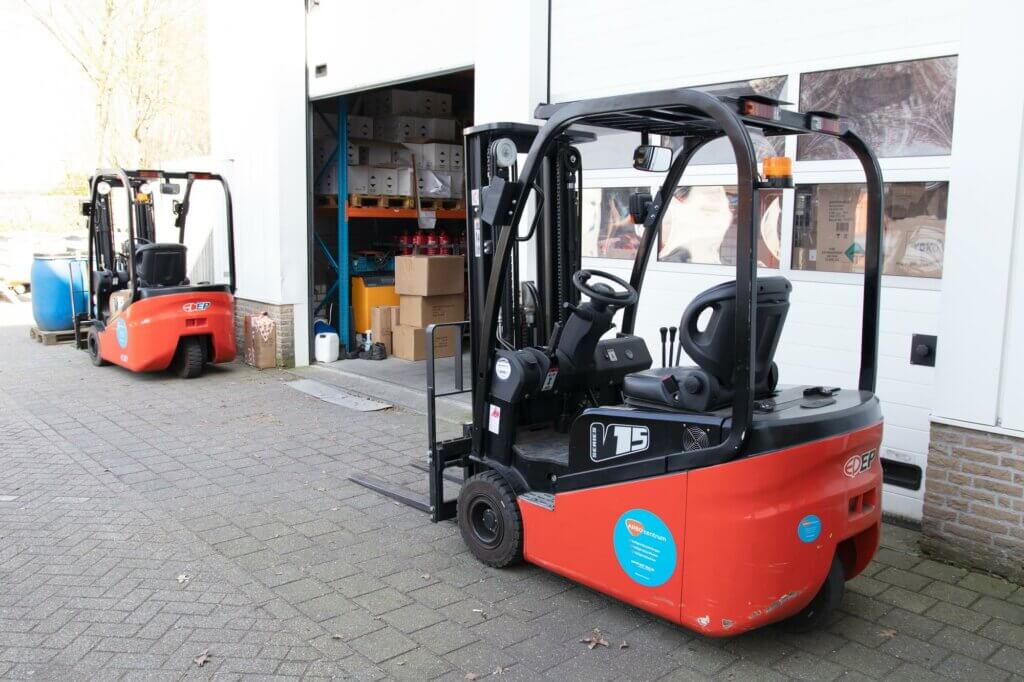 Twee rode en zwarte elektrische vorkheftrucks staan geparkeerd buiten een open magazijndeur. Eén staat op de voorgrond naar rechts gericht en de andere staat bij de ingang. Het behalen van een heftruckcertificaat biedt veiligere handling van dozen die op planken binnen zijn gestapeld, wat de praktische voordelen ervan laat zien.