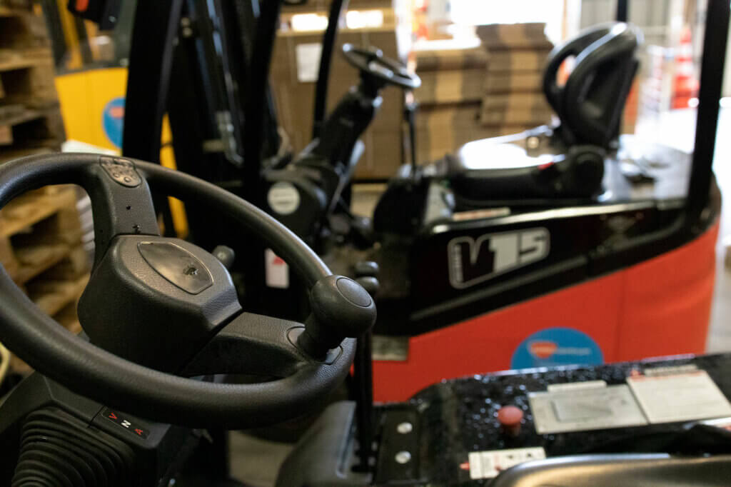 Close-up van een magazijnscène die 3 situaties laat zien waarbij je een heftruck gebruikt. De focus ligt op het stuur van een heftruck op de voorgrond, terwijl een andere op de achtergrond werkt. Stapels pallets en kartonnen dozen maken het tafereel compleet.