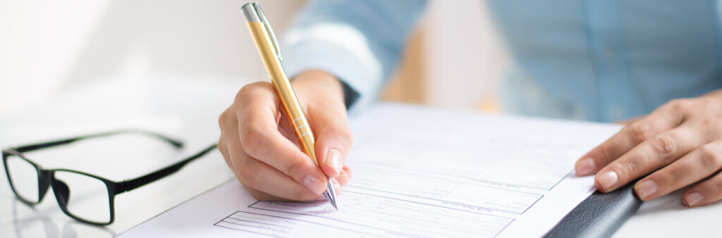 Een persoon in een blauw shirt schrijft met een pen op een document dat een speciale korting beschrijft, terwijl zijn bril op de tafel ernaast rust. De focus ligt op de hand en het papierwerk.