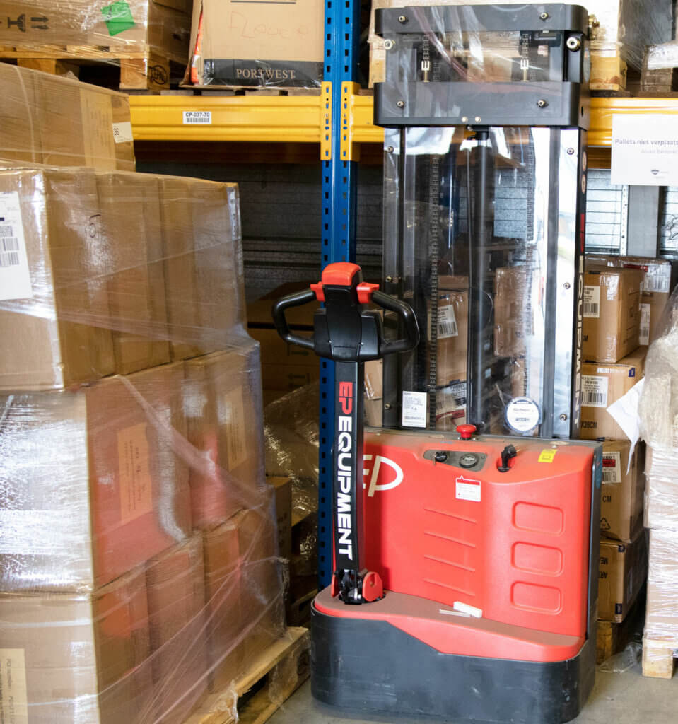 Een rode palletstapelaar met het merk EP Equipment staat geparkeerd in een gangpad in een magazijn, omringd door gestapelde kartonnen dozen en schappen. De stapelaar heeft een bedieningspaneel en een zwart beschermend frame, met praktische tips bij het besturen van een stapelaar voor een soepele werking.